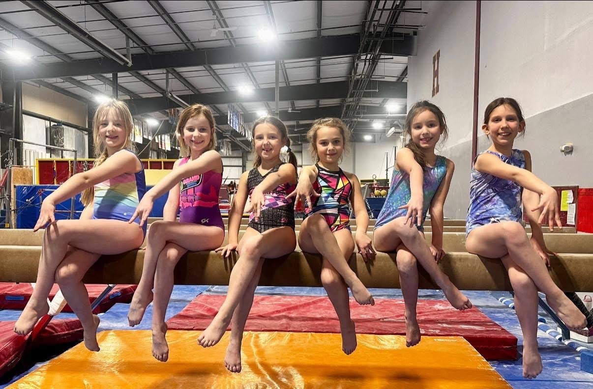 Six young gymnasts sitting on the balance beam at the gym